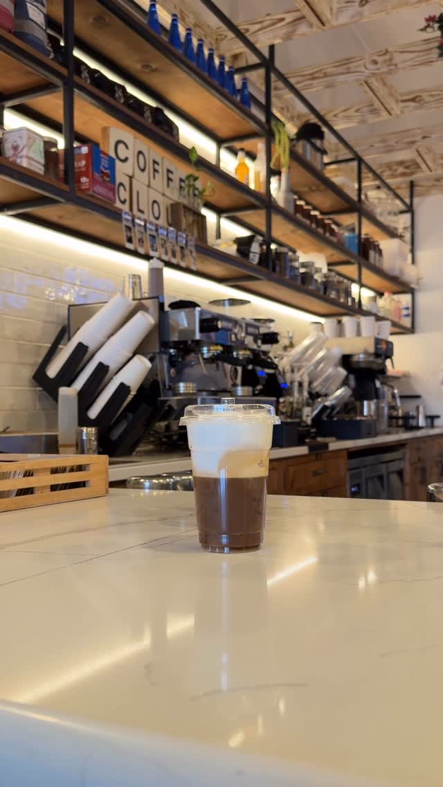 A freddo on the white-marble counter at Coffee Cloud, with the Scrabble-tile wordmark on the shelf
